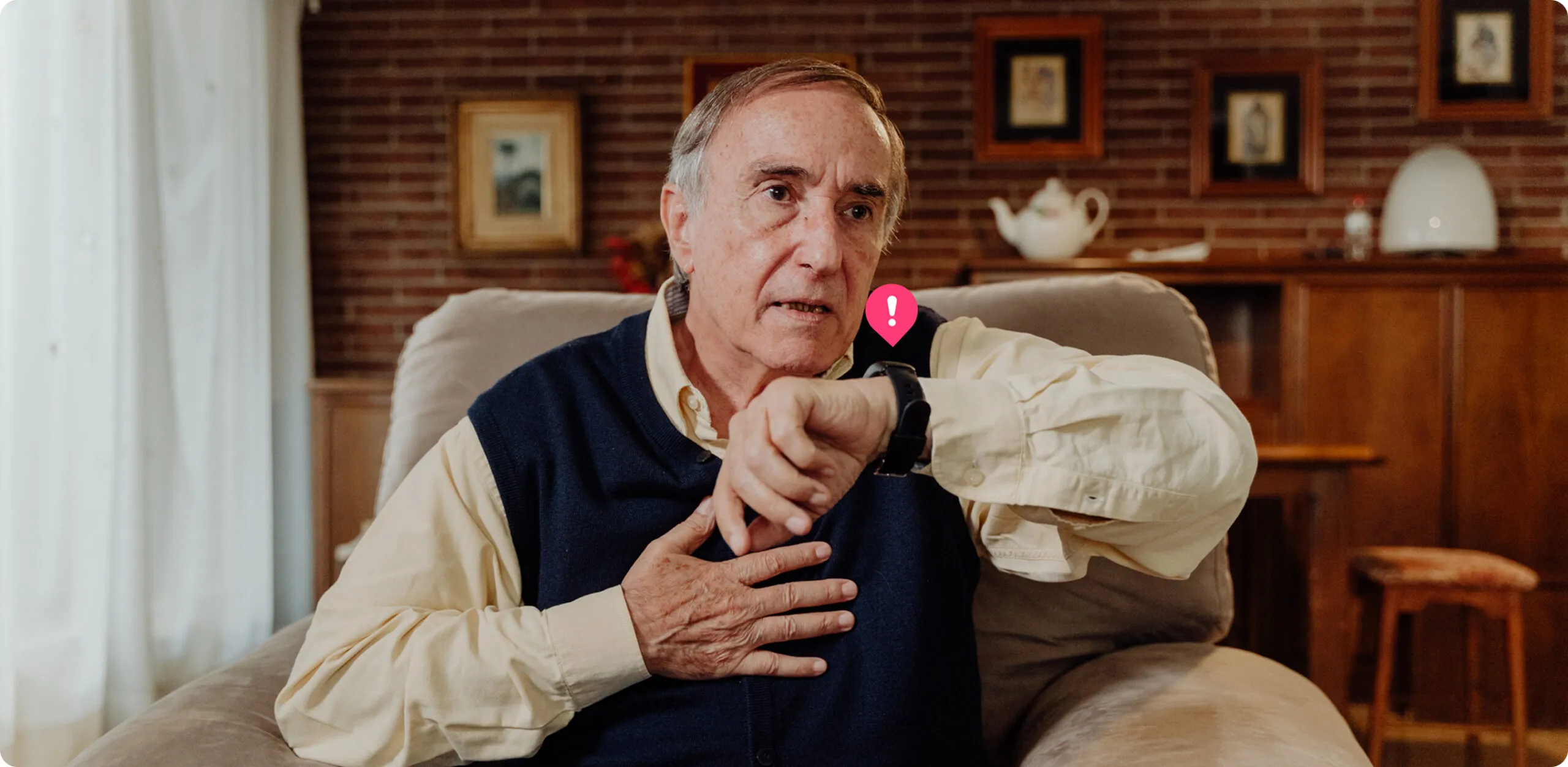 Un hombre sentado en un sillón pide ayuda a través de su reloj inteligente Durcal con expresión de preocupación, mientras mantiene una mano sobre su pecho. Sobre el reloj aparece un pequeño icono de notificación rosa.