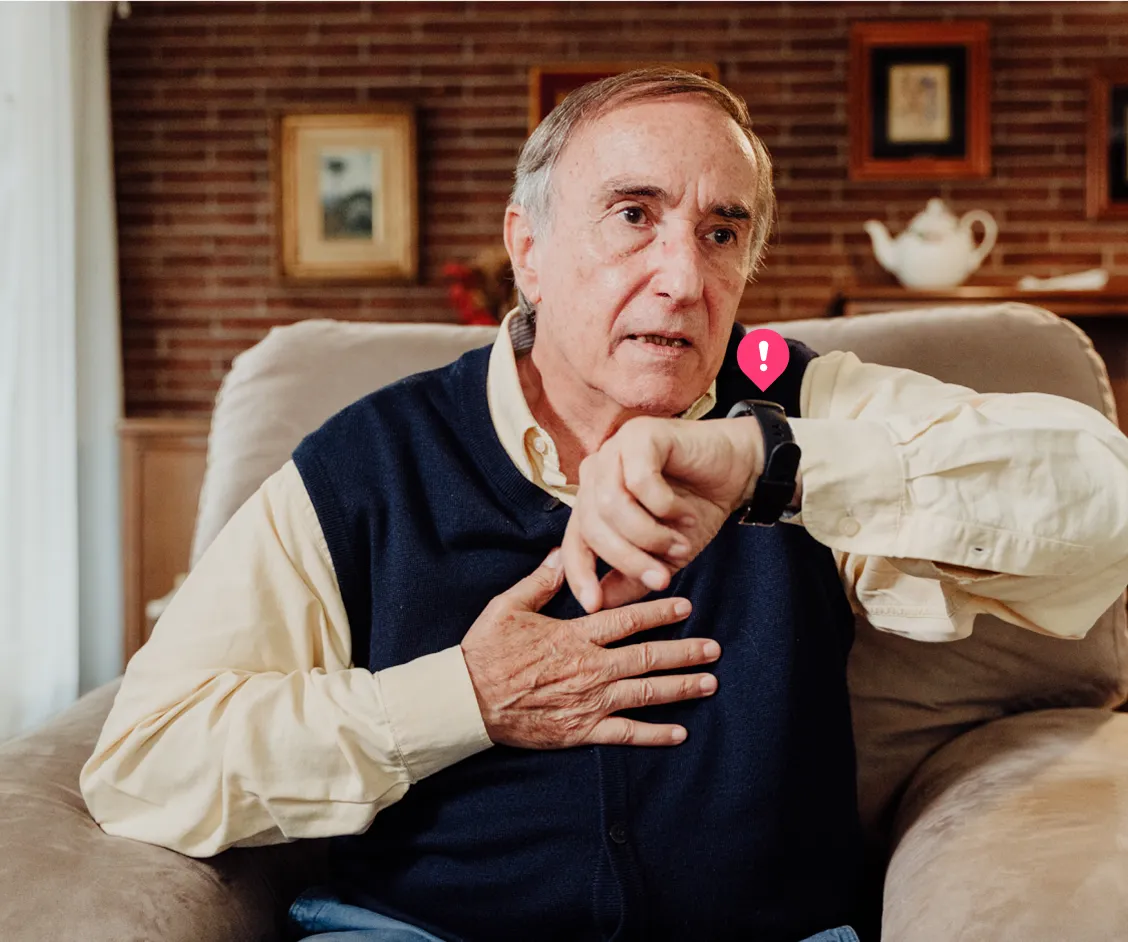 Un hombre sentado en un sillón pide ayuda a través de su reloj inteligente Durcal con expresión de preocupación, mientras mantiene una mano sobre su pecho. Sobre el reloj aparece un pequeño icono de notificación rosa.