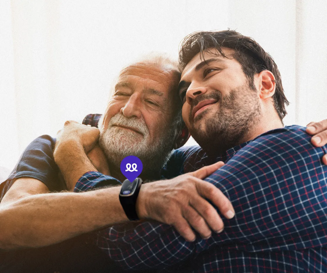 Un padre mayor con barba canosa y su hijo se abrazan felices y tranquilos en el salón de casa, reflejando paz mental gracias a la protección del reloj inteligente Durcal. En primer plano, destaca la muñeca del padre con el dispositivo negro y un icono morado flotante de la marca, simbolizando la seguridad de los mayores que viven solos y la tranquilidad de sus familias.