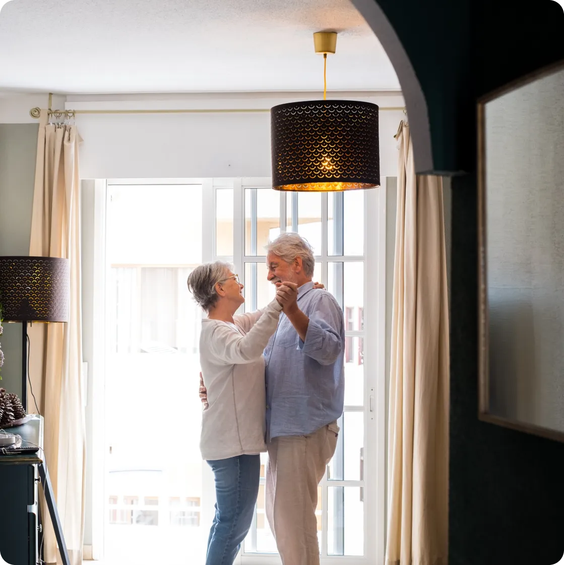 Una pareja de adultos mayores baila sonriente y abrazada en el salón de su casa, frente a un gran ventanal. La escena transmite alegría, autonomía y bienestar, reforzando la idea de que con la protección del reloj Durcal, las personas mayores pueden seguir disfrutando de sus momentos felices con total tranquilidad.