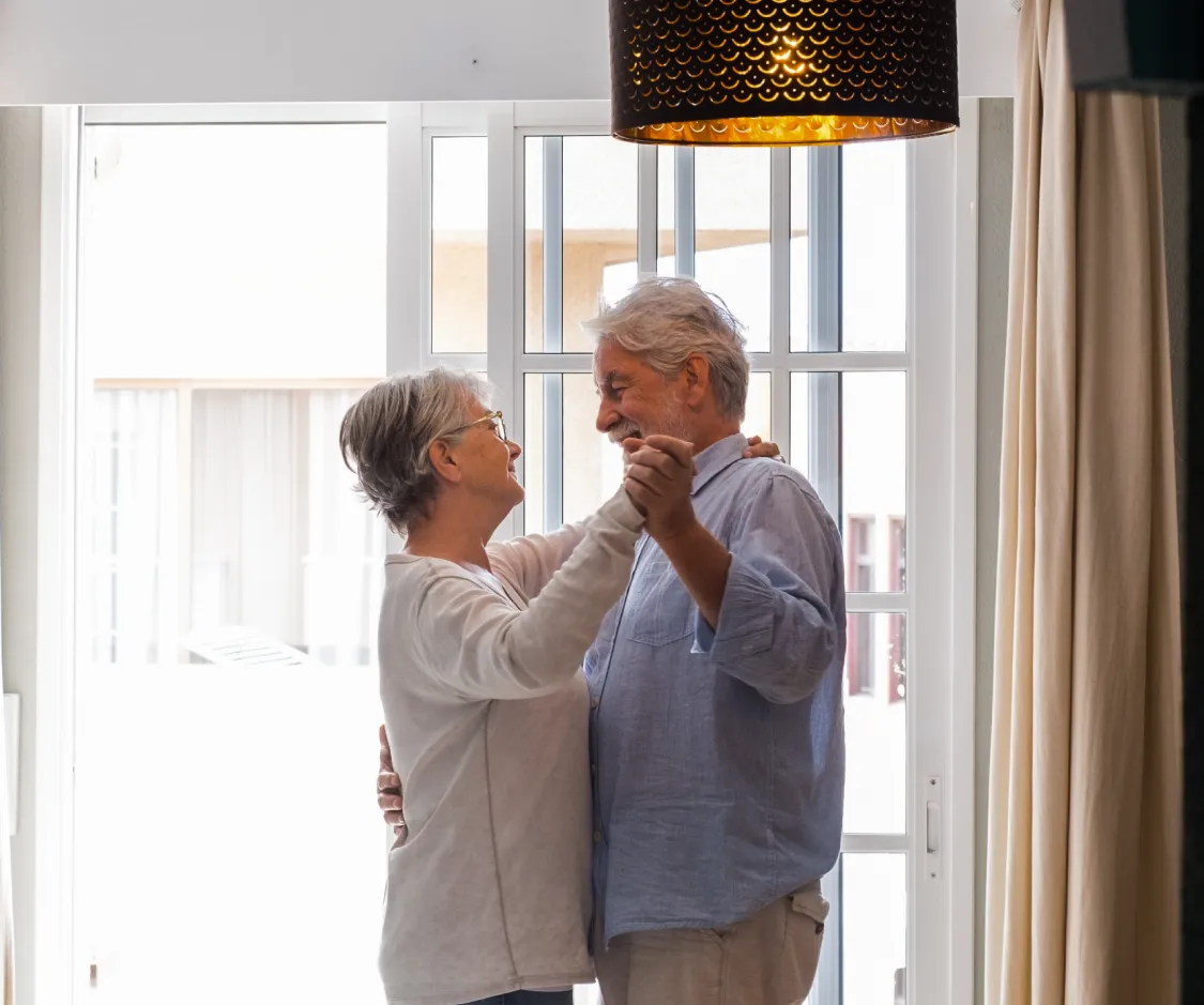 Una pareja de adultos mayores baila sonriente y abrazada en el salón de su casa, frente a un gran ventanal. La escena transmite alegría, autonomía y bienestar, reforzando la idea de que con la protección del reloj Durcal, las personas mayores pueden seguir disfrutando de sus momentos felices con total tranquilidad.