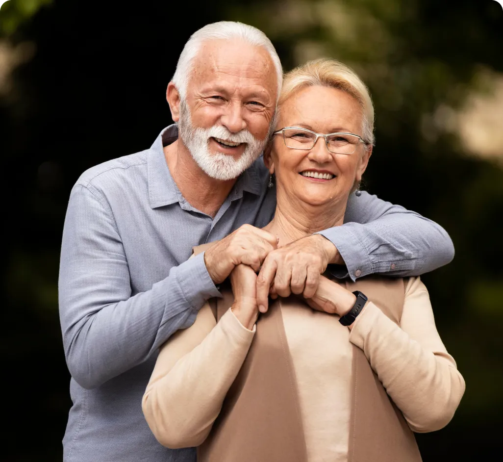 Una pareja de adultos mayores sonríe alegremente mientras el hombre abraza cariñosamente a la mujer por la espalda en un entorno exterior. La imagen transmite una sensación de vitalidad, seguridad y paz mental, reflejando cómo el respaldo de la teleasistencia de Durcal permite a los mayores y a sus familias disfrutar de la vida sin preocupaciones.