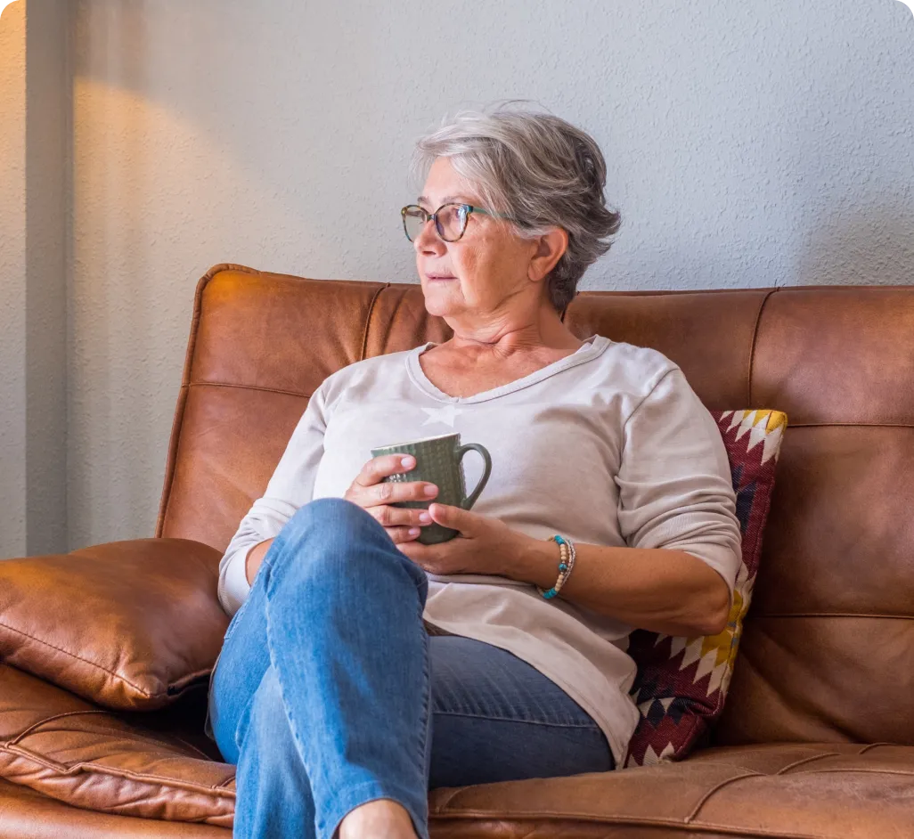 Una mujer mayor con gafas y cabello cano descansa tranquilamente en un sofá de cuero marrón, sosteniendo una taza entre sus manos y mirando hacia un lado con serenidad. La imagen evoca la calma y la independencia en el hogar, reforzada por la seguridad preventiva que ofrece el ecosistema de teleasistencia Durcal.