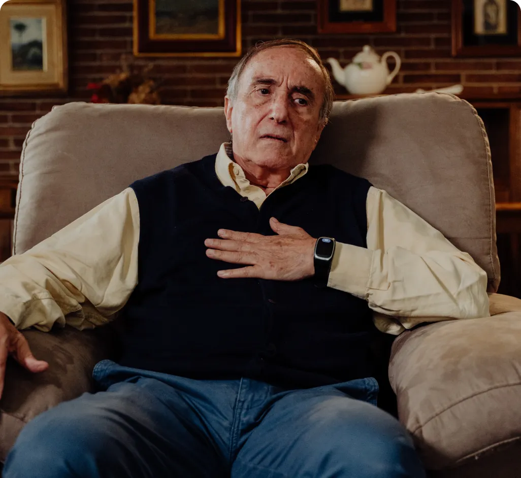 Un hombre mayor que vive solo se lleva la mano al pecho con gesto de malestar mientras descansa en un sillón, usando el reloj inteligente Durcal. La imagen representa la importancia de monitorizar la saturación de oxígeno y las constantes vitales en adultos mayores para prevenir complicaciones de salud de forma autónoma.