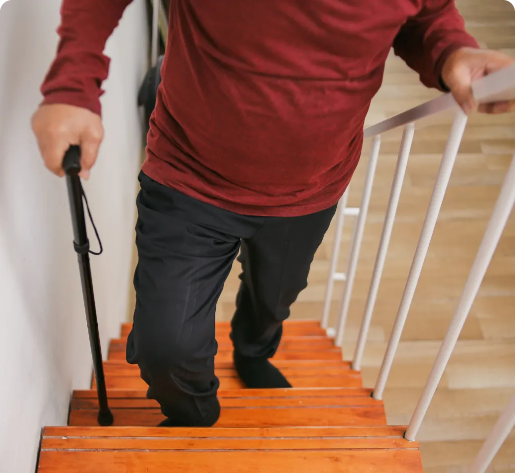 Vista frontal de un hombre mayor subiendo con esfuerzo unas escaleras de madera en su hogar, apoyándose en un bastón y en la barandilla. La imagen ilustra una situación cotidiana de movilidad reducida donde el reloj Durcal aporta seguridad ante el riesgo de caídas en el entorno doméstico.