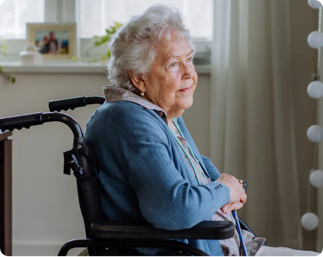 Una mujer mayor sentada en una silla de ruedas junto a una ventana luminosa en un entorno hogareño, mirando hacia el exterior con expresión tranquila.