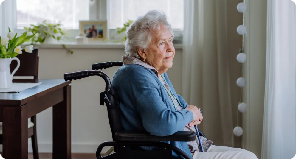 Una mujer mayor sentada en una silla de ruedas junto a una ventana luminosa en un entorno hogareño, mirando hacia el exterior con expresión tranquila.