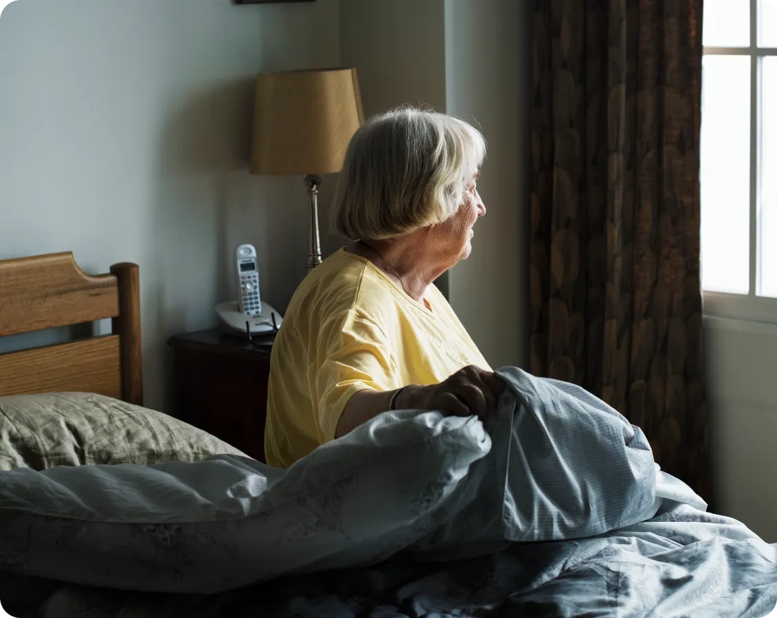 Una mujer mayor que vive sola se incorpora en su cama al amanecer, mirando pensativa hacia la ventana de su dormitorio. La escena subraya la importancia de contar con el reloj inteligente Durcal, que ofrece detección de caídas y ayuda inmediata, permitiendo que los adultos mayores mantengan su independencia y vivan solos en casa con total tranquilidad y conexión con sus familiares.