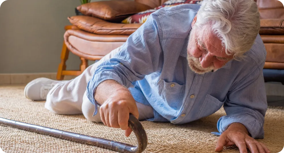 Un hombre mayor con el pelo canoso aparece tendido en la alfombra de su salón tras haber sufrido una caída, tratando de alcanzar su bastón. La imagen ilustra una situación de emergencia doméstica donde la función de detección de caídas del reloj Durcal permite enviar un aviso automático de ayuda.