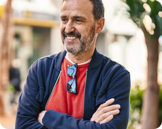 Retrato de medio cuerpo de un hombre con barba corta, sonriendo y con los brazos cruzados en un entorno exterior iluminado.