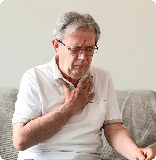 Un hombre mayor con gafas y cabello gris se sienta en un sofá y se lleva la mano al pecho con una expresión de dolor o malestar.