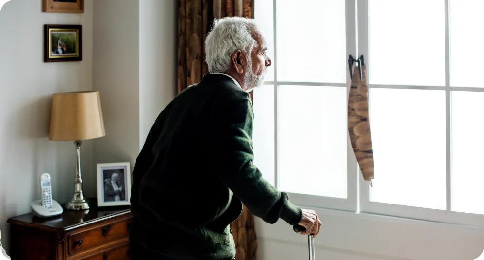 Un hombre mayor con barba y pelo canoso, vestido con un cárdigan verde, camina por su habitación apoyándose en un bastón mientras mira por la ventana. La imagen destaca la importancia de la autonomía en el hogar y cómo el reloj Durcal ofrece una red de seguridad invisible para quienes viven solos o tienen movilidad reducida.