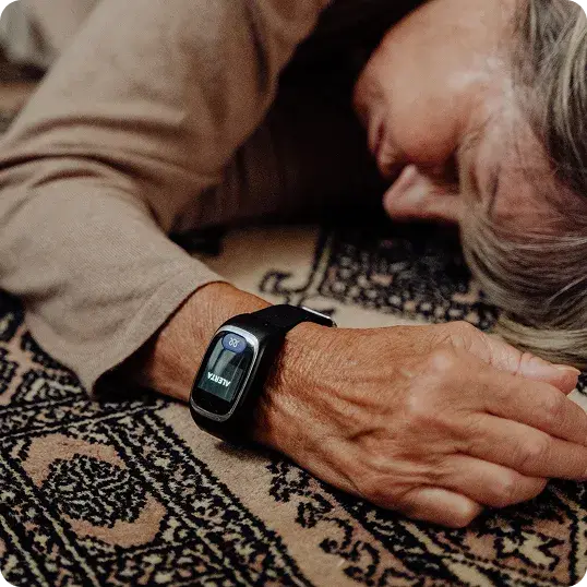Una mujer mayor tirada en el suelo sobre una alfombra estampada tiene un smartwatch negro en su muñeca izquierda. La pantalla del reloj muestra la palabra 'ALERTA'