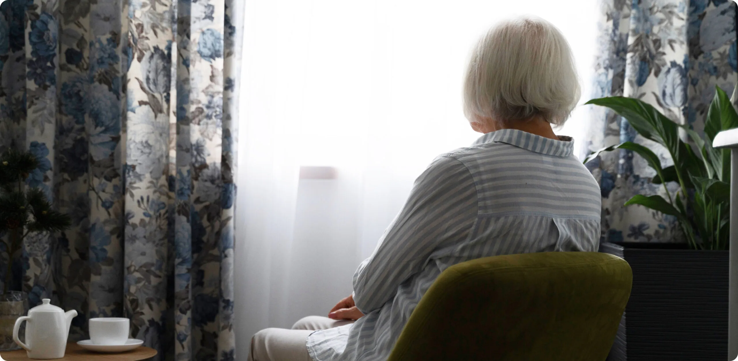 Una mujer mayor con cabello cano vista de espaldas, sentada en un sillón verde y mirando por una ventana luminosa en su hogar. La imagen transmite una sensación de soledad o espera, sugiriendo la importancia de la teleasistencia de Durcal para mantener conectados y seguros a los adultos mayores que viven solos.