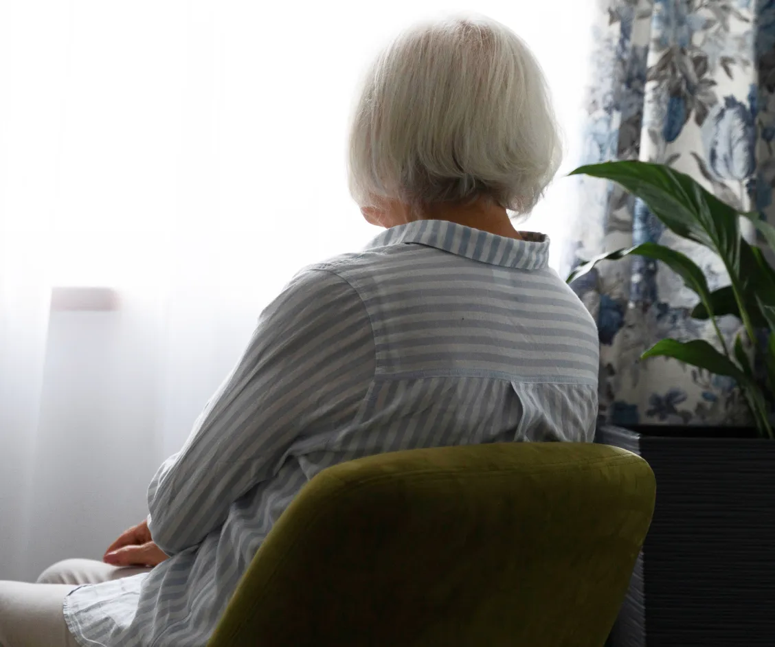 Una mujer mayor con cabello cano vista de espaldas, sentada en un sillón verde y mirando por una ventana luminosa en su hogar. La imagen transmite una sensación de soledad o espera, sugiriendo la importancia de la teleasistencia de Durcal para mantener conectados y seguros a los adultos mayores que viven solos.