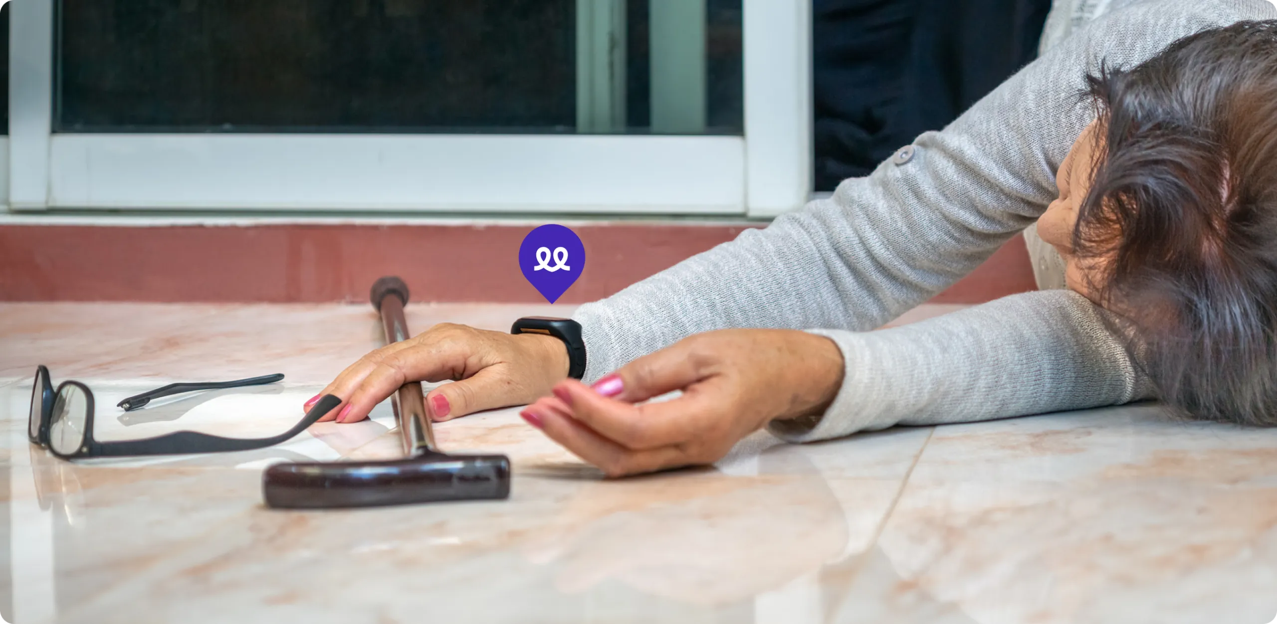Primer plano de una persona mayor tendida en el suelo tras una caída, con un bastón a su lado. En su muñeca lleva el reloj Durcal, resaltado por un icono de ubicación morado, simbolizando la función de detección de caídas y aviso automático a emergencias o familiares.