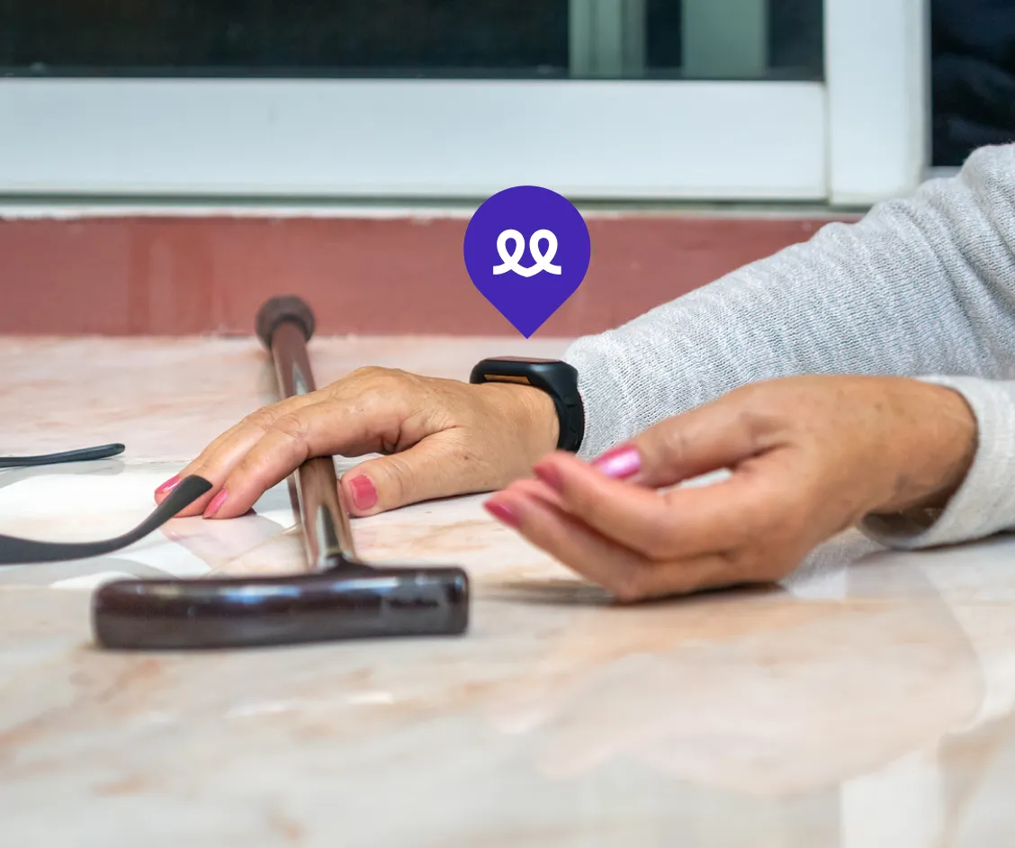 Primer plano de una persona mayor tendida en el suelo tras una caída, con un bastón a su lado. En su muñeca lleva el reloj Durcal, resaltado por un icono de ubicación morado, simbolizando la función de detección de caídas y aviso automático a emergencias o familiares.