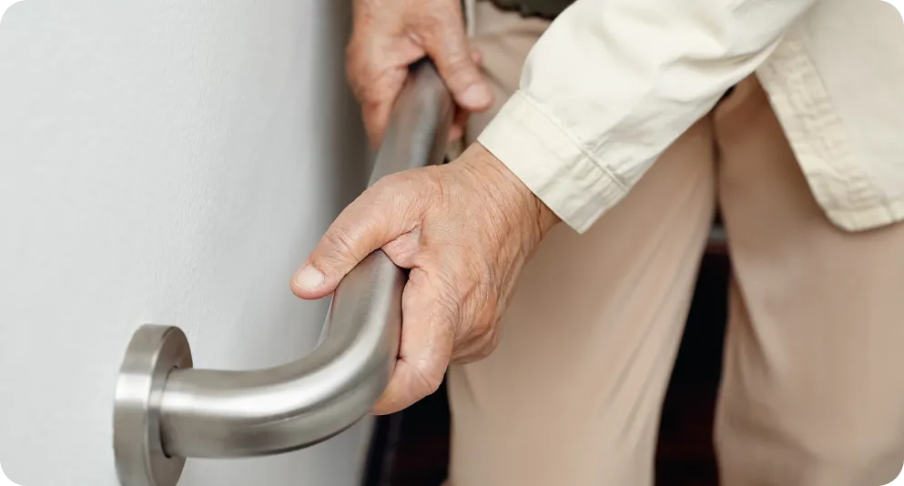 Primer plano de las manos de una persona mayor sujetándose con firmeza a un asidero de seguridad metálico en la pared. La imagen representa las medidas de prevención en el hogar y la importancia de contar con el respaldo de la teleasistencia de Durcal ante posibles problemas de equilibrio o movilidad.